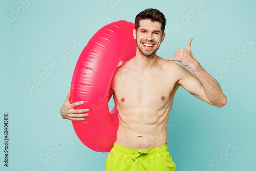 Young sexy man wear green shorts swimsuit relax near hotel pool hold inflatable rubber ring show shaka sign look camera isolated on plain blue cyan background Summer vacation sea rest sun tan concept photo