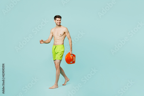 Full body young lifeguard sexy man wear green shorts swimsuit relax near hotel pool hold lifebuoy walk go look aside on area isolated on plain blue background Summer vacation sea rest sun tan concept