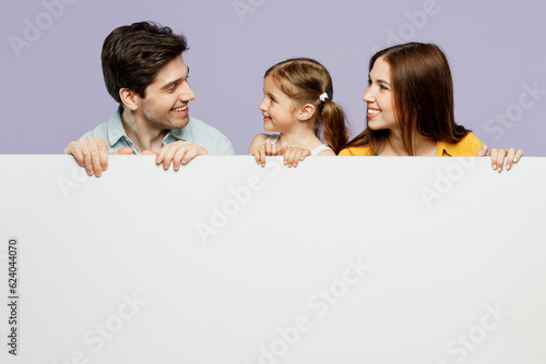 Young amazed parents mom dad with child kid daughter girl 6 years old wearing blue yellow casual clothes hold big white empty poster billboard isolated on plain purple background. Family day concept.