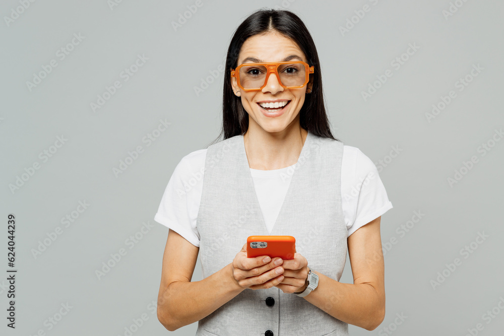 Young happy smiling latin woman wear white t-shirt gray vest glasses hold in hand use mobile cell phone in red case chatting online isolated on plain grey background studio portrait Lifestyle concept Young happy smiling latin woman wear white t-shirt gray vest glasses hold in hand use mobile cell phone in red case chatting online isolated on plain grey background studio portrait Lifestyle concept