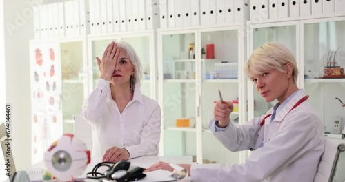 Female doctor points to Snellen chart to check patient vision. Doctor sitting at clinic workplace with model of eyeball examines elderly lady patient vision photo