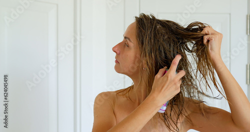 Young woman in towel near mirror using hair spray in bedroom