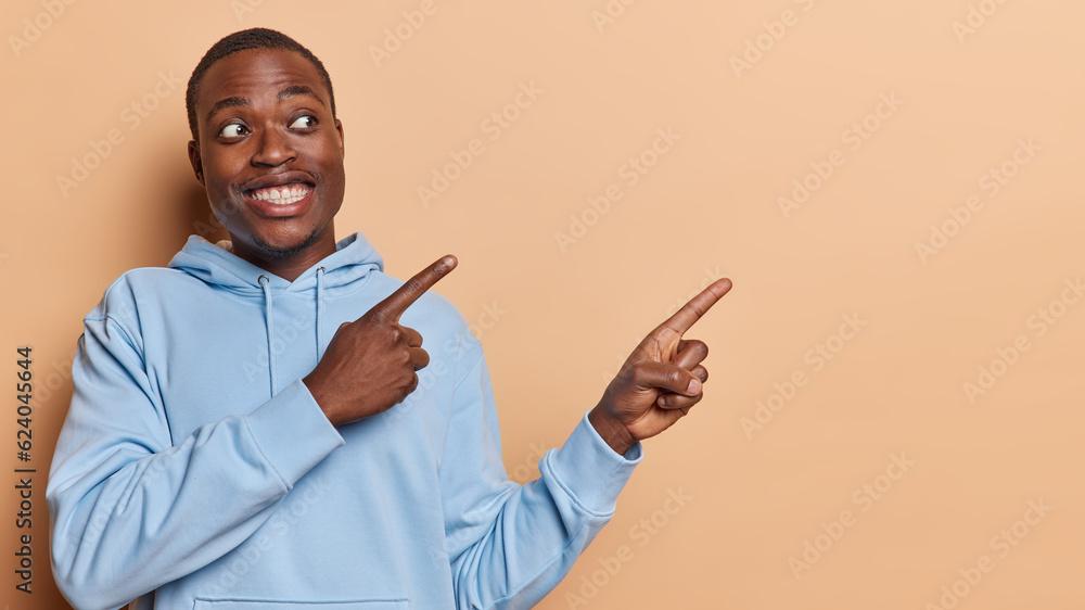 Handsome dark skinned man points index fingers aside on blank space shows advertisement or promo smiles broadly shows white teeth dressed in casual blue sweatshirt isolated over brown background Handsome dark skinned man points index fingers aside on blank space shows advertisement or promo smiles broadly shows white teeth dressed in casual blue sweatshirt isolated over brown background