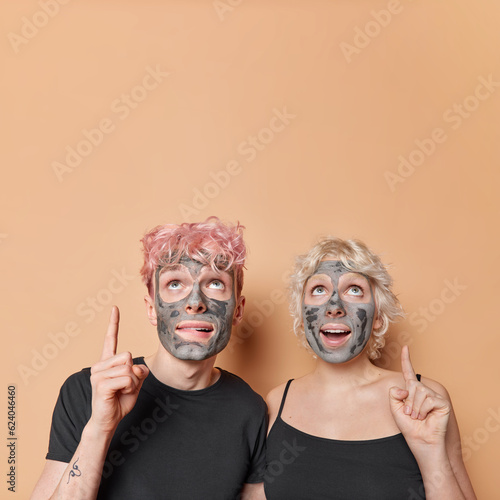 Vertical shot of positive woman and man apply facial clay mask for skin treatment indulge in shared moment of self care point index fingers overhead isolated on brown background copy space for advert
