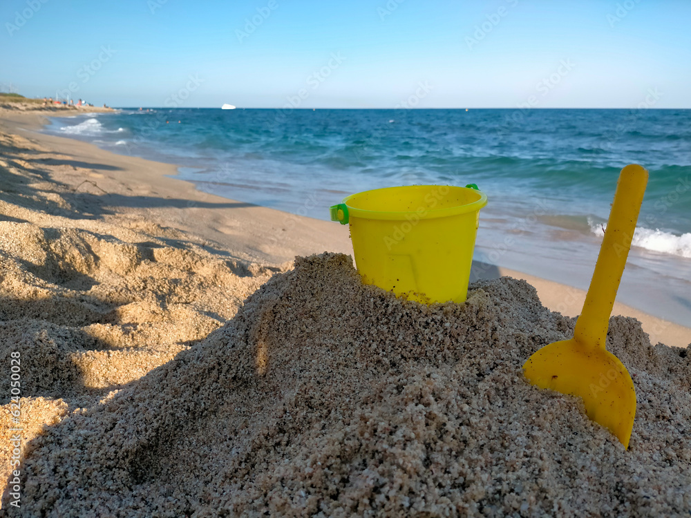 a yellow bucket and shovel in the sand on the beach for children to play sand castles, you can also see the Mediterranean Sea and the beach a yellow bucket and shovel in the sand on the beach for children to play sand castles, you can also see the Mediterranean Sea and the beach