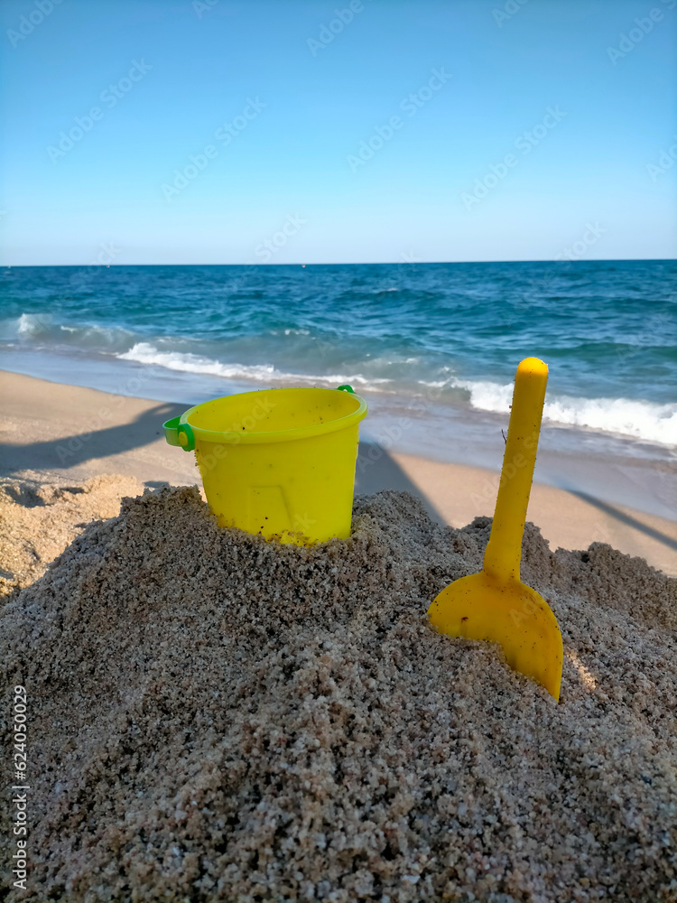 a yellow bucket and shovel in the sand on the beach for children to play sand castles, you can also see the Mediterranean Sea and the beach a yellow bucket and shovel in the sand on the beach for children to play sand castles, you can also see the Mediterranean Sea and the beach