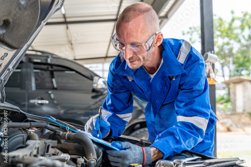 A male auto mechanic in blue jumpsuit writing engine repair notes on his paper notepad while working its engine at car repair garage