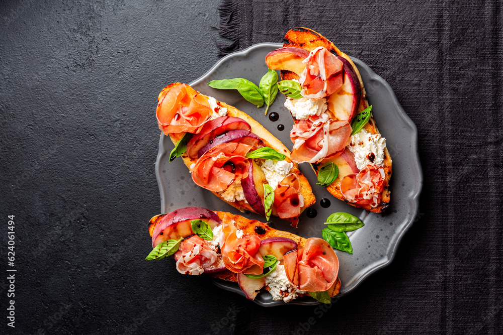 A dark grey plate with with open sandwiches, snack. Toastet bread, griddled peach, parma ham or prosciuto and robiola cheese, with balsamic vinegar and basil. Black background, top view. A dark grey plate with with open sandwiches, snack. Toastet bread, griddled peach, parma ham or prosciuto and robiola cheese, with balsamic vinegar and basil. Black background, top view.