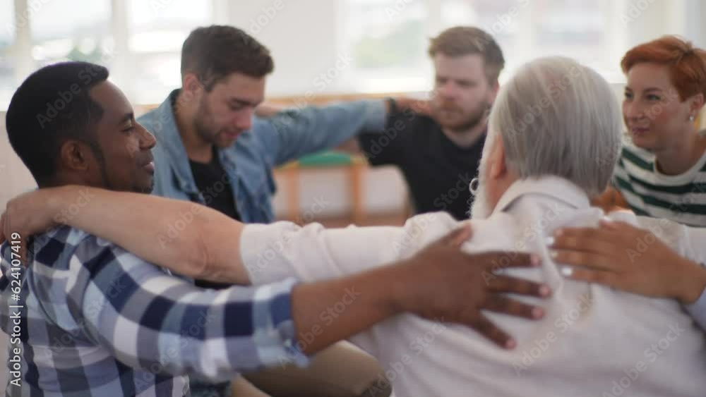 Back view of mature male therapist and group of multi-age patients putting hands on each others shoulders sitting in circle during group therapy session as symbol of support. Shooting in slow motion. Back view of mature male therapist and group of multi-age patients putting hands on each others shoulders sitting in circle during group therapy session as symbol of support. Shooting in slow motion.