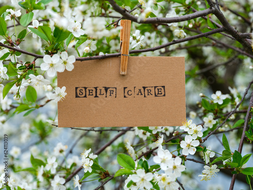 A piece of cardboard with the word Self Care on it hanging on a cherry tree branch with blossoms using a wooden clothes pin. © rosinka79