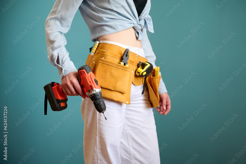 Young female worker with tool belt holding an electric screwdriver in hands isolated on background. Pretty caucasian female with tools planning new project. Portrait in studio. Crop view. Young female worker with tool belt holding an electric screwdriver in hands isolated on background. Pretty caucasian female with tools planning new project. Portrait in studio. Crop view.