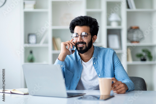 Happy Indian Male Having Phone Conversation And Drinking Coffee At Home Office