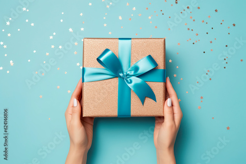  First person top view photo of hands unpacking craft paper gift box with blue satin ribbon bow over shiny sequins on isolated pastel blue background