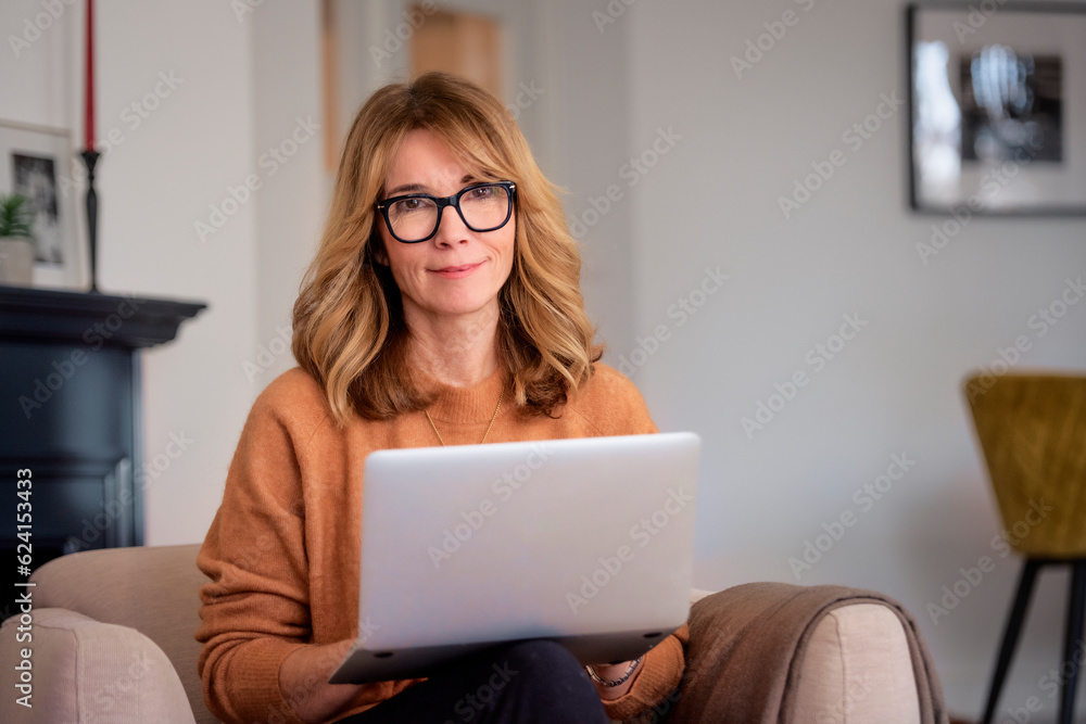 Attractive middle aged woman sitting in an armchair at home and using laptop for work Attractive middle aged woman sitting in an armchair at home and using laptop for work