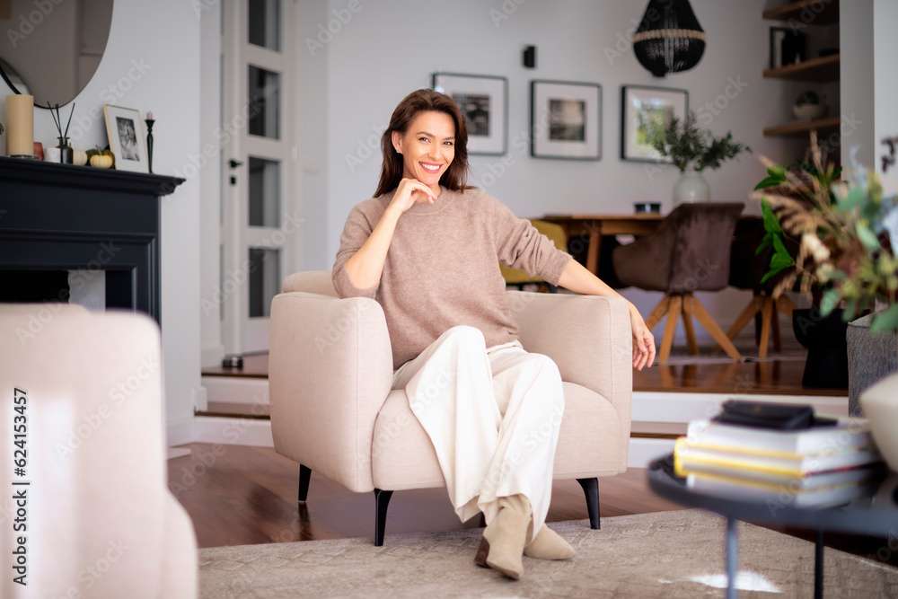 Obraz premium Attractive mid aged woman wearing sweater and white pants and sitting in an armchair in her modern home