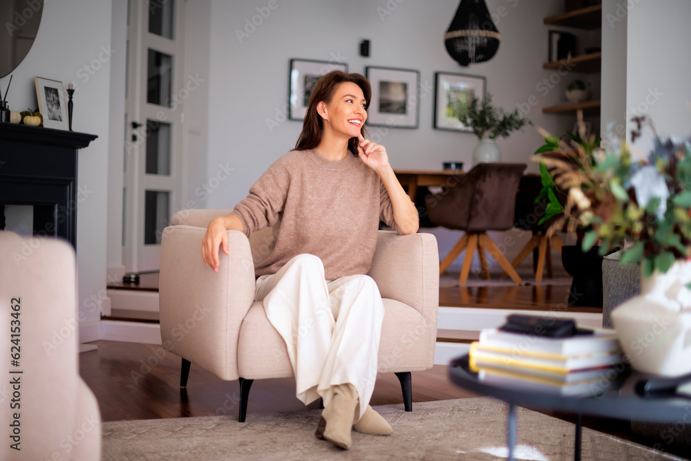 Attractive mid aged woman wearing sweater and white pants and sitting in an armchair in her modern home