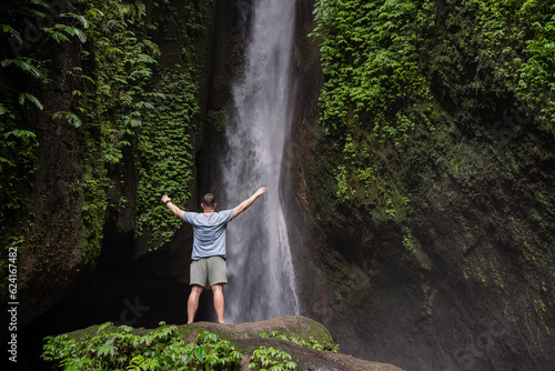 Back view of a man standing by waterfalls with arms outstretched. Man at waterfall raising his hands in feeling closer to nature. Man at the waterfall. Travel to Bali