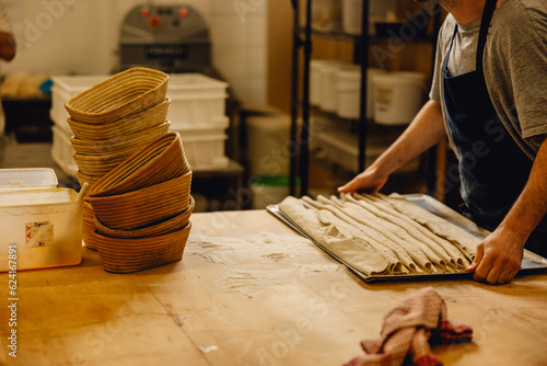 baker lifts tray of proving baguettes off wooden table in professional bakery kitchen with stack of proving baskets nearby