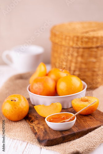 Close-up of plum jam in a white bowl and yellow plums whole and slices on a wooden board in a cozy bright kitchen, vertical photo.