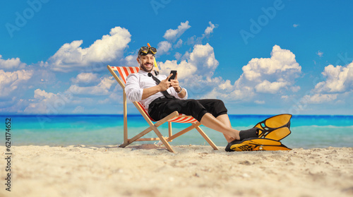 Businessman with snorkelling fins and mask sitting on a bech chair by the sea and using a mobile phone