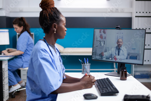 Medical team on internet videoconference telehealth video call in busy clinical office. African american nurse at hospital desk consulting with general practitioner about patient case