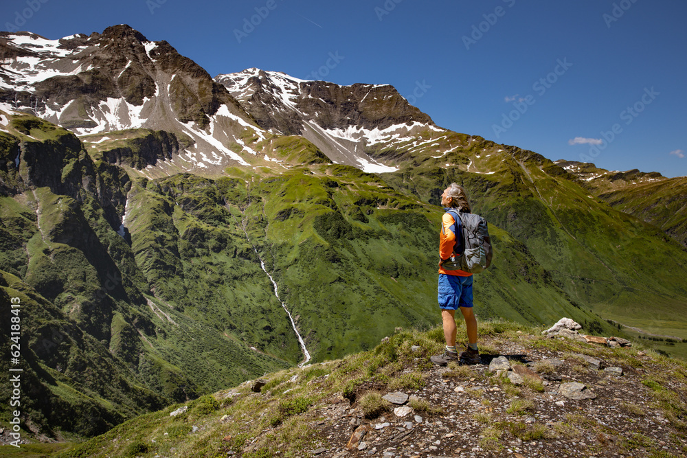 A gray-haired man stands on mountain path and admires the mountain landscape with green slopes and snowy peaks. Austria