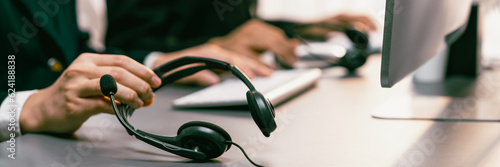 Panorama focus hand holding headset on call center workspace desk with blur background of operator team or telesales representative engaging in providing client with customer support service. Prodigy