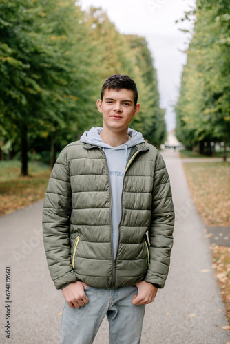 a guy in a jacket stands on an alley in the park during the fall season. Portrait oh handsome teen guy, young man in hoodie, down jacket standing, walking in beautiful golden autumn park, looking at © andreiko