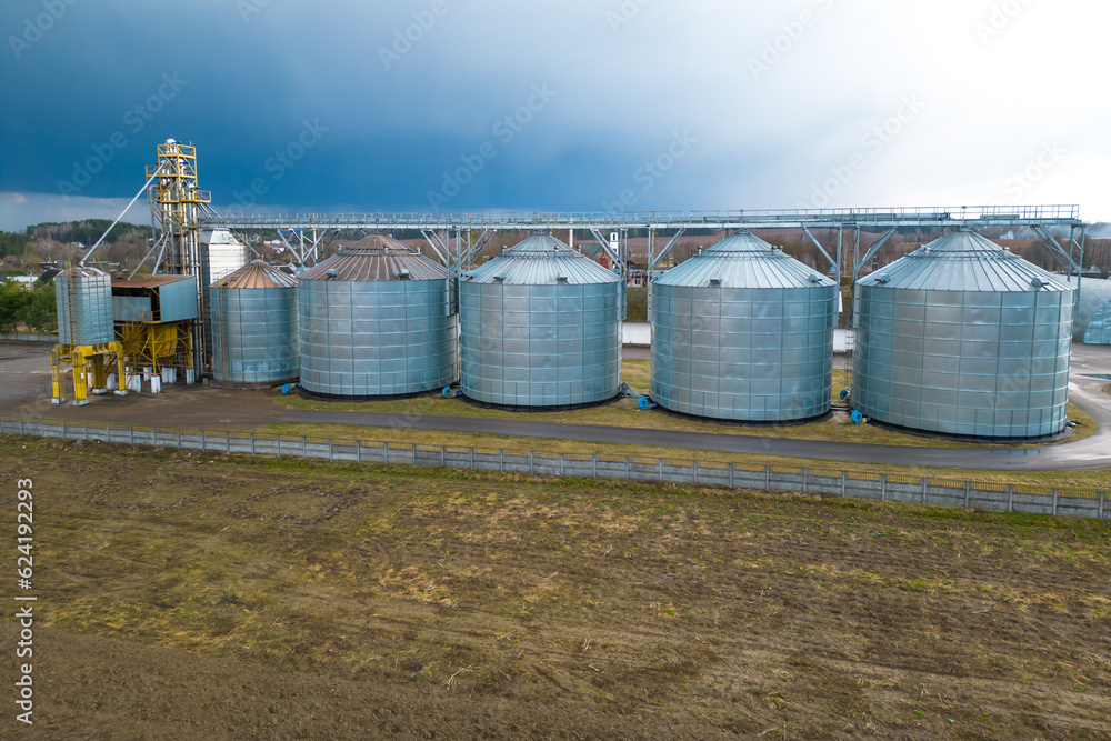 Flying a drone over silver silos on agro manufacturing plant for processing drying cleaning and storage of agricultural products, flour, cereals and grain. Large iron barrels of grain. aerial view Flying a drone over silver silos on agro manufacturing plant for processing drying cleaning and storage of agricultural products, flour, cereals and grain. Large iron barrels of grain. aerial view