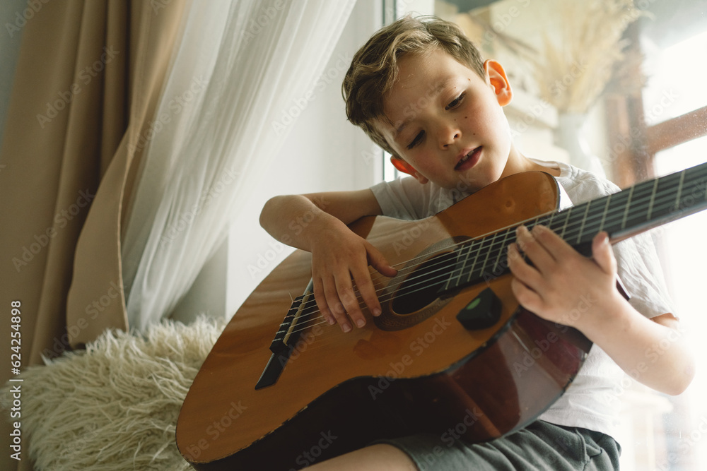 Fototapeta premium Cute boy learns to play the classical guitar on the windowsill near the window. Cozy home. Summer holidays lifestyle.