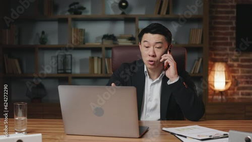 Busy office employee having serious conversation with business clients on smartphone while working on portable computer. Good-looking asian man sitting in modern workplace and solving issues remotely.