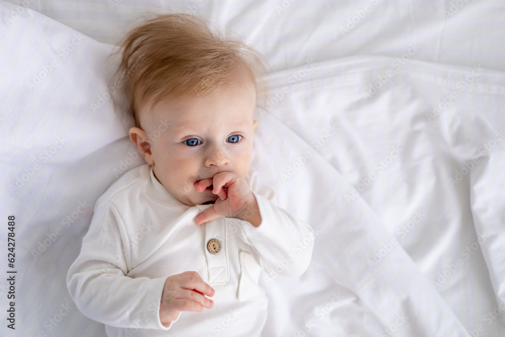 smiling baby 6 months old blond boy lies on a white bed in a bright bedroom and licks his finger in a cotton bodysuit, the concept of children's goods smiling baby 6 months old blond boy lies on a white bed in a bright bedroom and licks his finger in a cotton bodysuit, the concept of children's goods