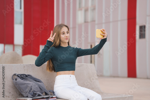 Young teenage student having fun taking a selfie sitting on a university campus. Excited Caucasian looking at the camera showing the peace sign outdoors.
