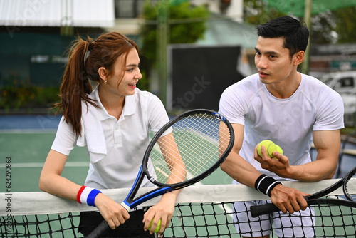 Experienced male tennis coach giving instructions to his student, standing by net at the outdoor tennis court © Prathankarnpap