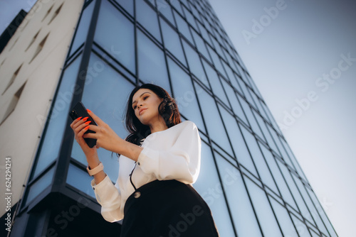 Young cheerful business woman in stylish suit standing outside in sunny day and looking with toothy smile at screen of modern smartphone in hand, chatting with friend or writing message to colleague.