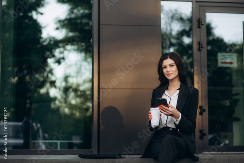 Young cheerful business woman in stylish suit standing outside in sunny day and looking with toothy smile at camera with modern smartphone in hand, chatting with friend or writing message to colleague