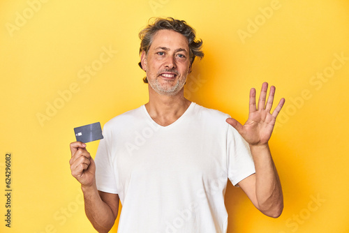 Man holding a credit card, yellow studio backdrop smiling cheerful showing number five with fingers.