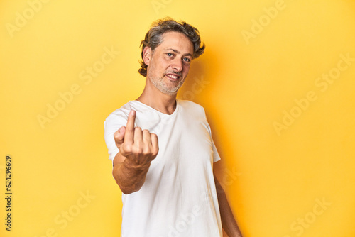 Middle-aged man posing on a yellow backdrop pointing with finger at you as if inviting come closer. © Asier