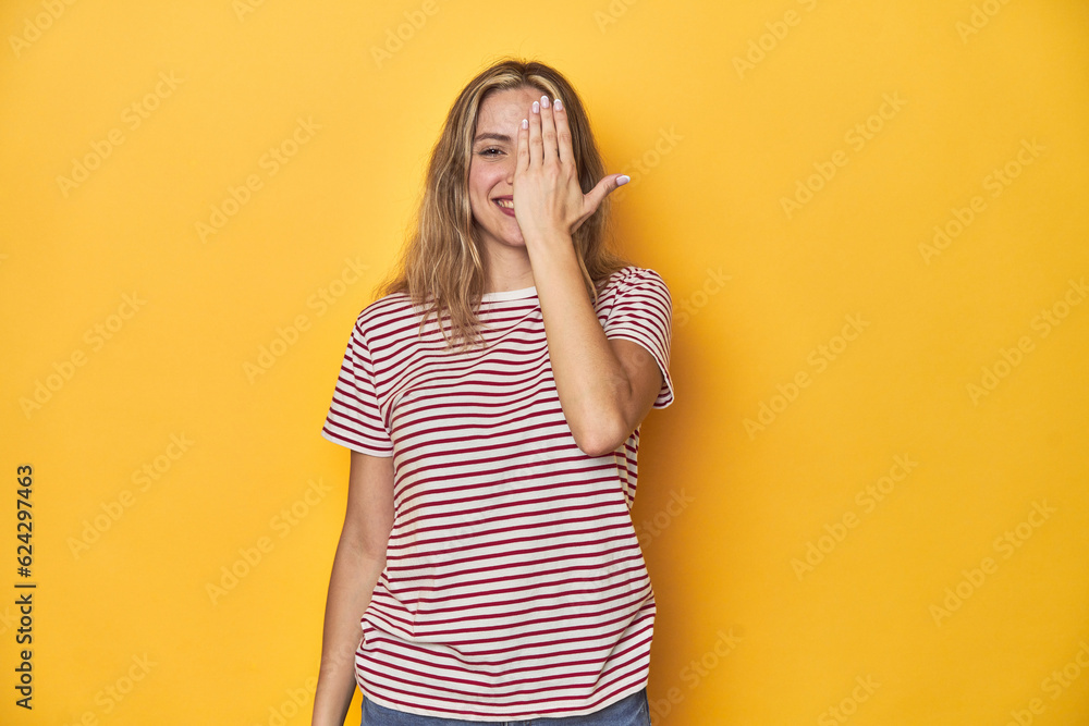 Young blonde Caucasian woman in a red striped t-shirt on a yellow background, having fun covering half of face with palm. Young blonde Caucasian woman in a red striped t-shirt on a yellow background, having fun covering half of face with palm.