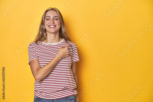 Young blonde Caucasian woman in a red striped t-shirt on a yellow background, smiling and pointing aside, showing something at blank space.