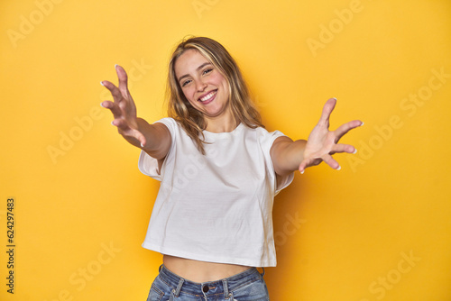 Young blonde Caucasian woman in a white t-shirt on a yellow studio background, feels confident giving a hug to the camera.