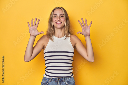 Young blonde Caucasian woman in a white tank top on a yellow studio background, showing number ten with hands.