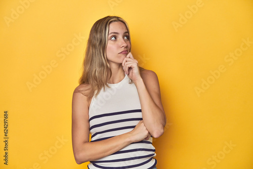 Young blonde Caucasian woman in a white tank top on a yellow studio background, thinking and looking up, being reflective, contemplating, having a fantasy.
