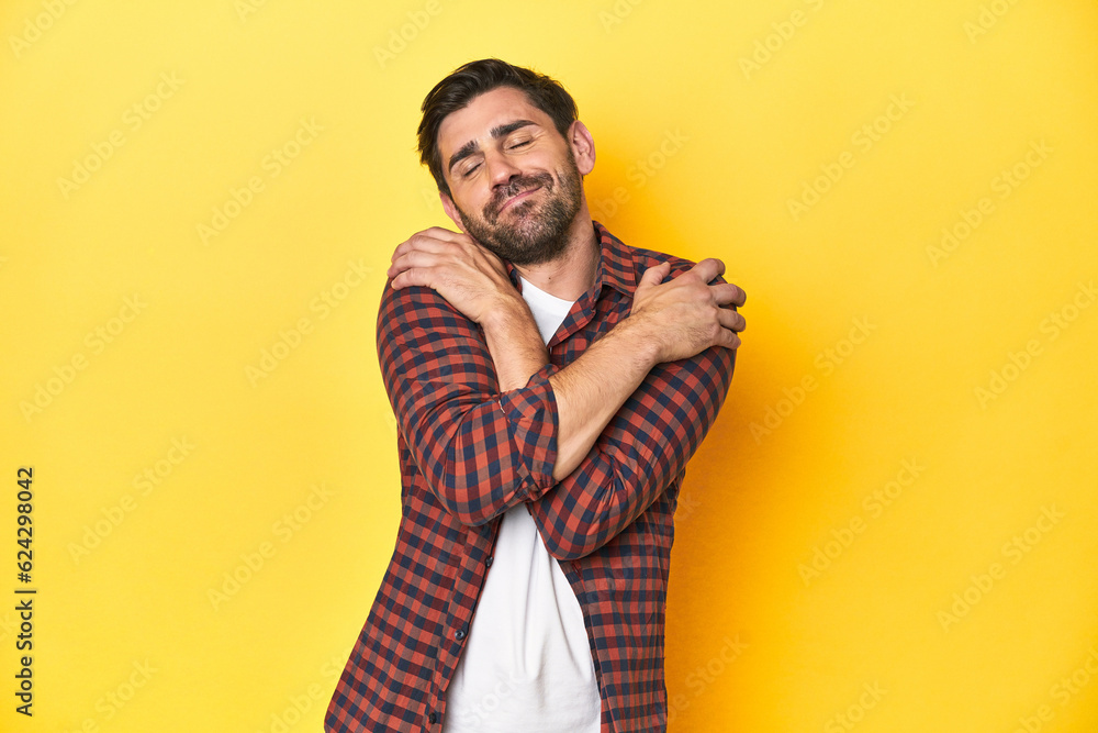 Fototapeta premium Caucasian man in red checkered shirt, yellow backdrop hugs, smiling carefree and happy.