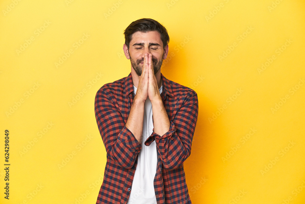 Caucasian man in red checkered shirt, yellow backdrop holding hands in pray near mouth, feels confident. Caucasian man in red checkered shirt, yellow backdrop holding hands in pray near mouth, feels confident.