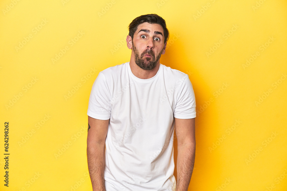 Caucasian man in white t-shirt on yellow studio background shrugs shoulders and open eyes confused. Caucasian man in white t-shirt on yellow studio background shrugs shoulders and open eyes confused.