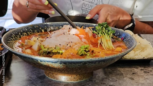 Woman eating Mulhoe raw fish cold soup inside korean restaurant. Close-up woman's hands picking up fish slices with vegetables from plate in slow motion photo