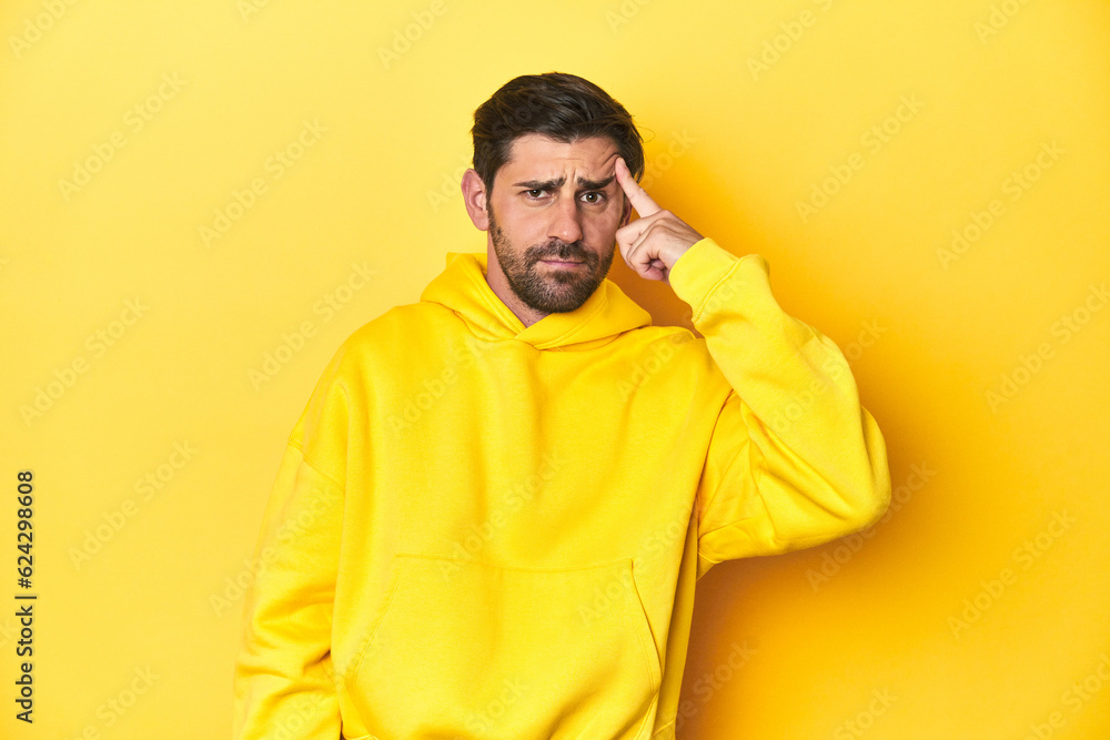 Man in yellow hoodie, monochrome studio backdrop pointing temple with finger, thinking, focused on a task. Man in yellow hoodie, monochrome studio backdrop pointing temple with finger, thinking, focused on a task.