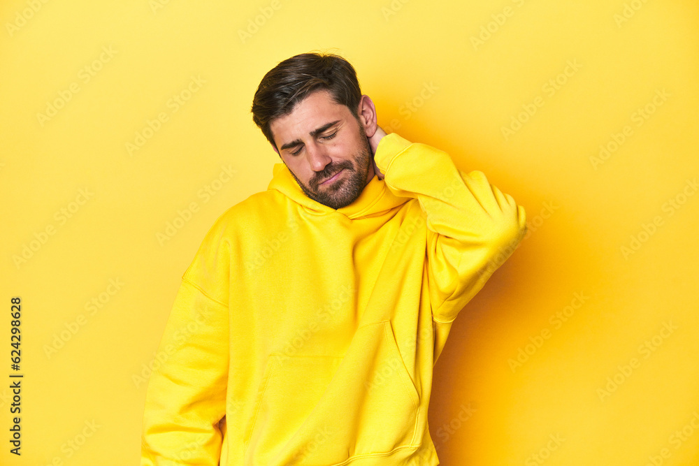 Man in yellow hoodie, monochrome studio backdrop suffering neck pain due to sedentary lifestyle. Man in yellow hoodie, monochrome studio backdrop suffering neck pain due to sedentary lifestyle.