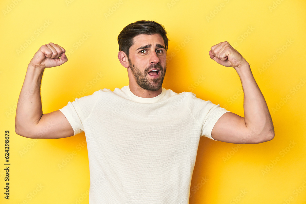 Caucasian man in white t-shirt on yellow studio background showing strength gesture with arms, symbol of feminine power Caucasian man in white t-shirt on yellow studio background showing strength gesture with arms, symbol of feminine power
